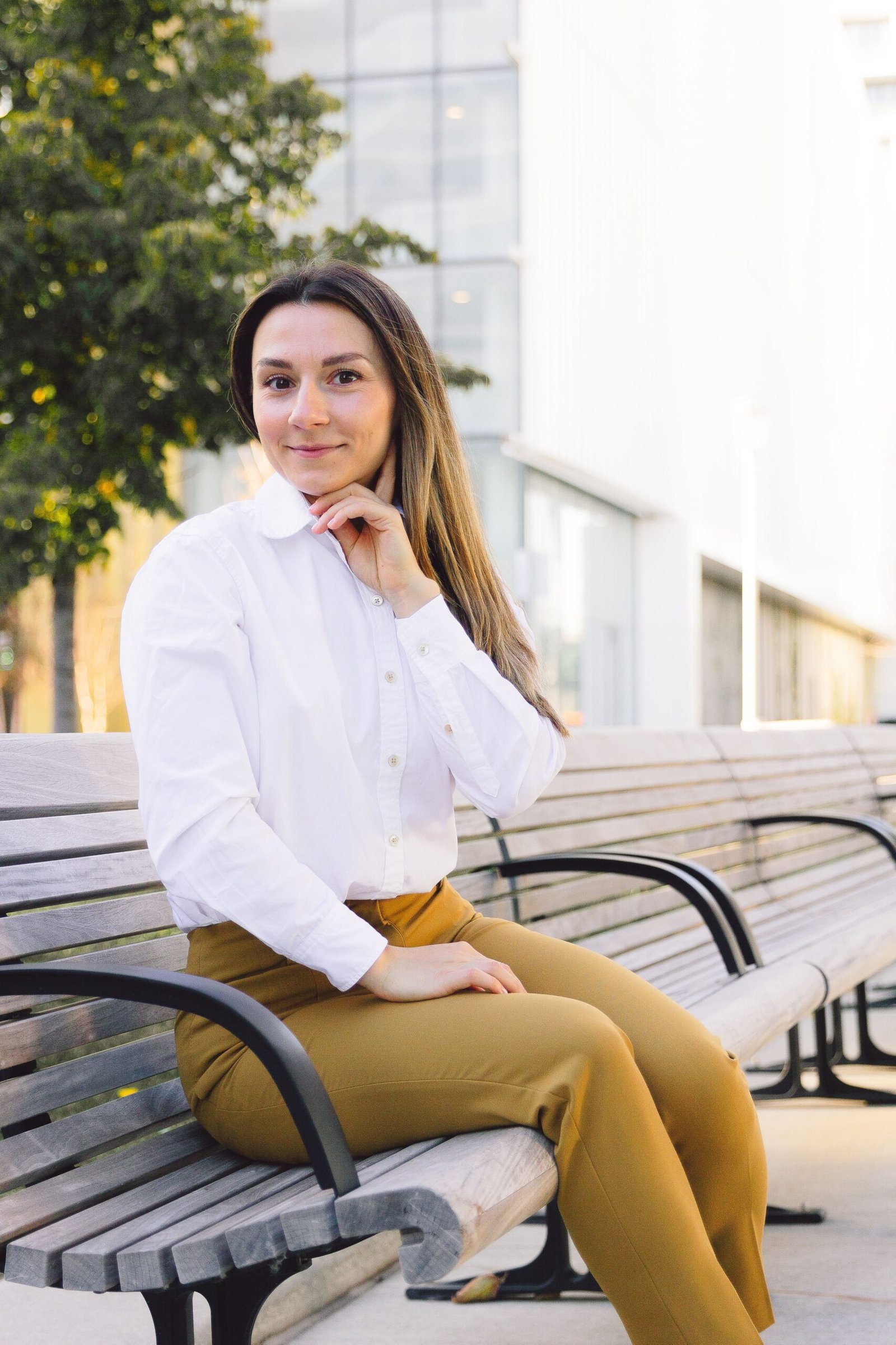 Young woman in business casual attire sitting on an outdoor bench in a city setting.